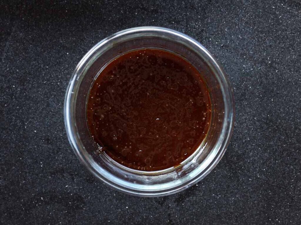 A glass bowl filled with dark brown sauce sits on a black textured countertop. The sauce appears thick and glossy, with a consistent texture throughout the bowl.